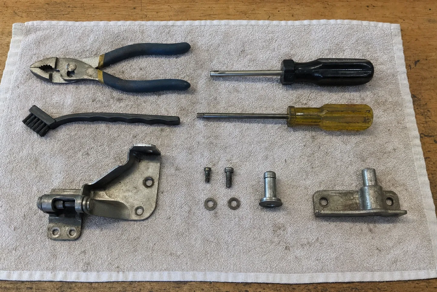 Door hinge assembly with its pivot pin, screws and washers laid out on a cloth beside pliers, two screwdrivers and a wire brush