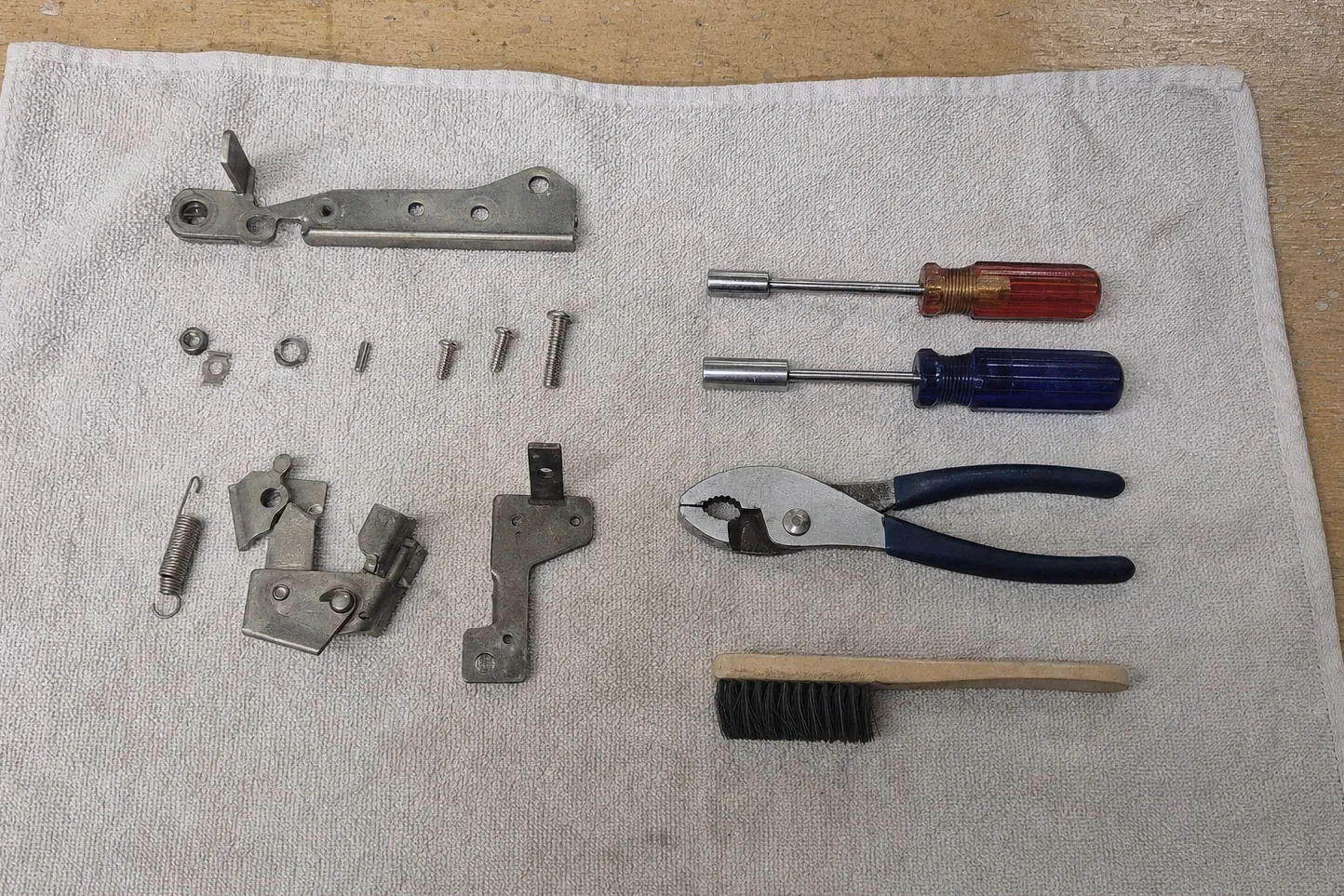 Door latch and hinge parts with a return spring, small screws, nuts and washers laid out on a cloth beside two nut drivers and pliers
