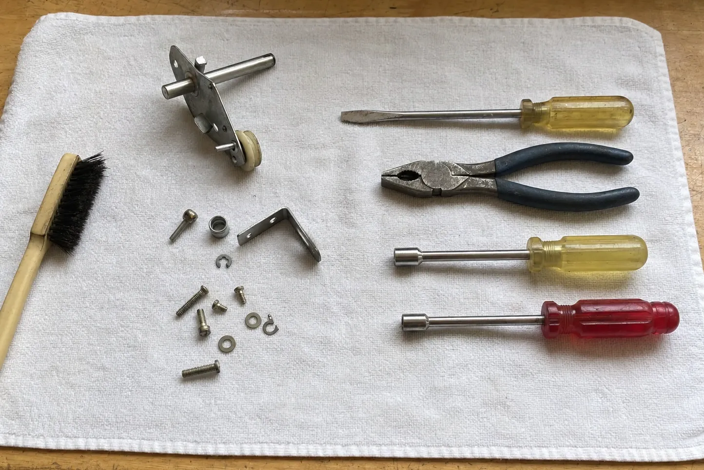 Hinge and roller assembly with its nylon roller, an angle bracket, a clip and fasteners on a cloth beside nut drivers and pliers