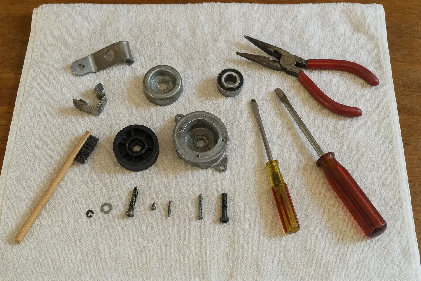 Sealed ball bearing, a cast bearing housing with its cover off, a black nylon wheel and an alloy roller set out on a towel with a spring clip, screws, a wire brush, screwdrivers and needle-nose pliers. Numbered marks are drawn over it, at the sealed ball bearing, bearing housing with the cover off and the nylon wheel.