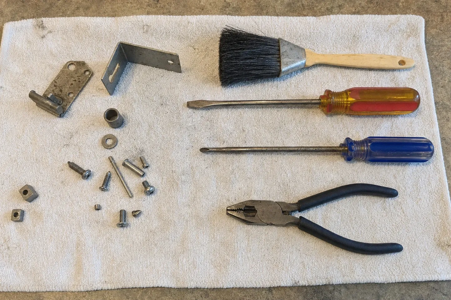Flat steel plate carrying a pivot barrel and a stub arm, an L-bracket with a keyhole slot and a sleeve bushing on a towel with bolts, square nuts, a dusting brush, screwdrivers and pliers. Numbered marks are drawn over it, at the keyhole slot in the bracket, pivot barrel on the plate and the sleeve bushing.