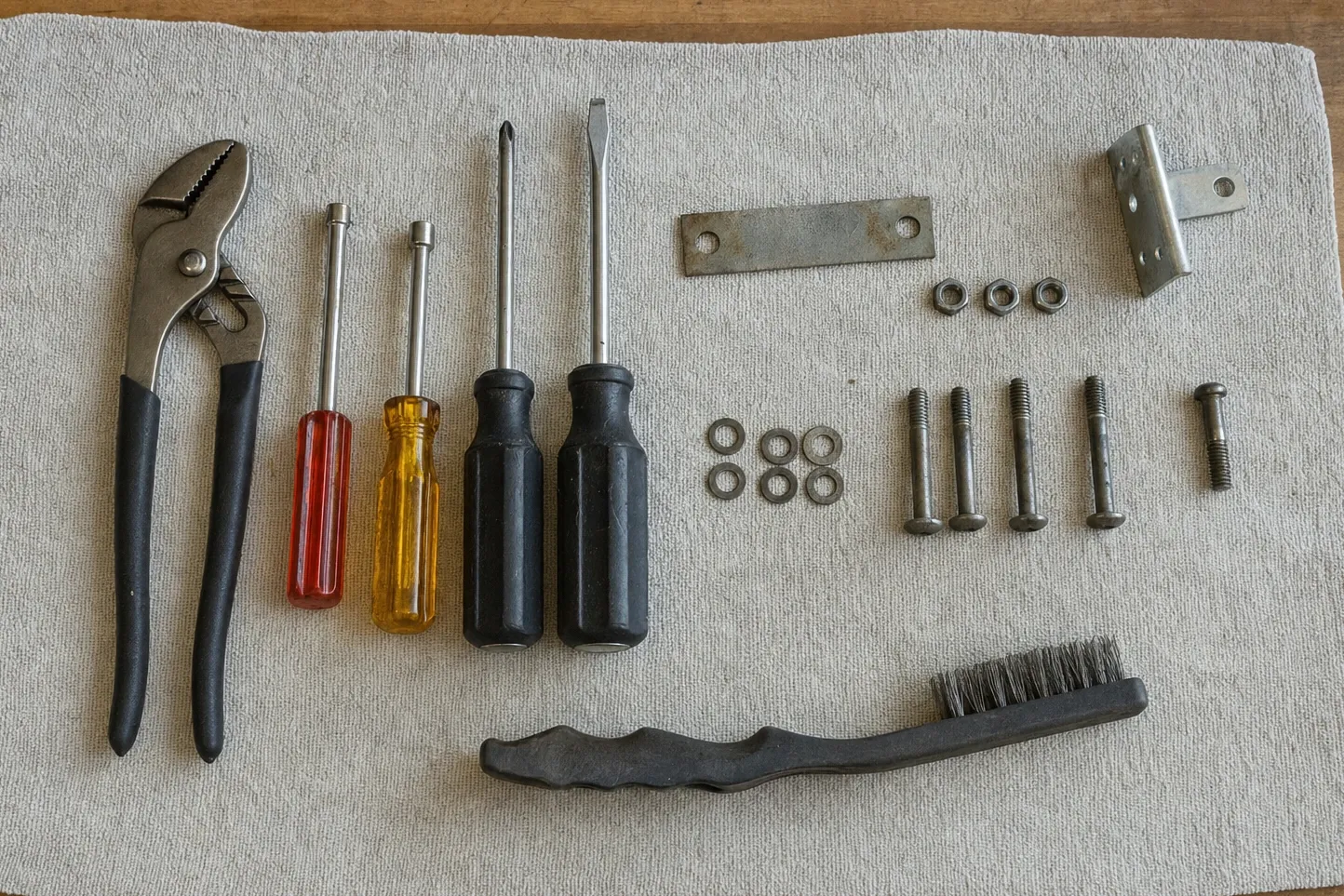Flat two-hole strap plate and an L-bracket with hex nuts, six washers and five machine screws in rows on a linen cloth beside tongue-and-groove pliers, nut drivers, screwdrivers and a wire brush. Numbered marks are drawn over it, at the flat two-hole strap plate, l-bracket and the machine screws and washers.